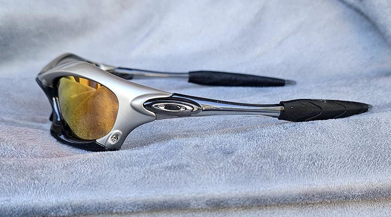Może przedstawiać: Srebrno-czarne okulary przeciwsłoneczne sportowe ze złotymi soczewkami. Okulary przeciwsłoneczne mają elegancki, nowoczesny design z zakrzywioną oprawką i czarnymi gumowymi końc&oacute;wkami zausznik&oacute;w. Soczewki są w ciepłym, złotym kolorze.