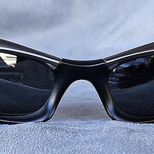 Peut inclure: Lunettes de soleil noires avec un design incurv&eacute; et enveloppant. Les verres sombres refl&egrave;tent l'environnement. Les montures sont noires mates, pos&eacute;es sur un tissu bleu. Un accessoire de lunetterie moderne.