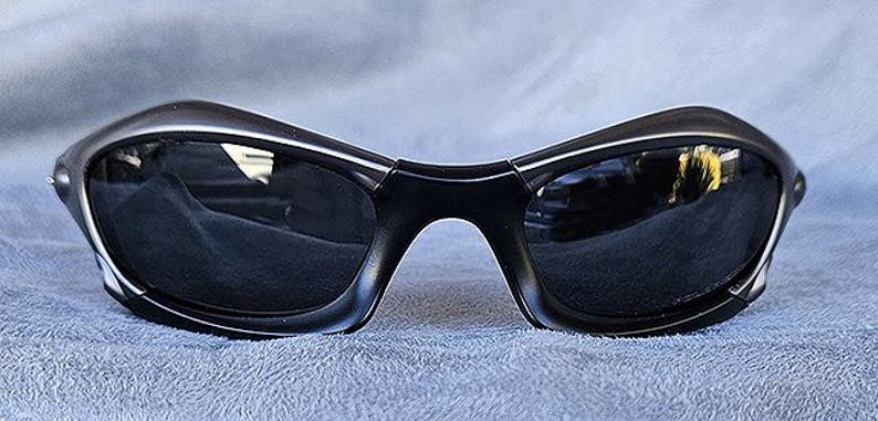 Peut inclure: Lunettes de soleil noires avec un design incurv&eacute; et enveloppant. Les verres sombres refl&egrave;tent l'environnement. Les montures sont noires mates, pos&eacute;es sur un tissu bleu. Un accessoire de lunetterie moderne.