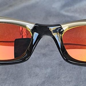 Może przedstawiać: Sportowe okulary przeciwsłoneczne ze złotą oprawką i czarnymi akcentami. Soczewki mają pomarańczowo-ż&oacute;łty gradient. Okulary mają zakrzywioną konstrukcję i są przeznaczone do aktywności na świeżym powietrzu.
