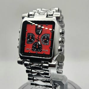 May include: A silver-toned Oakley watch with a square red face. The watch features multiple black dials and a silver metal band. The watch is displayed on a reflective surface.
