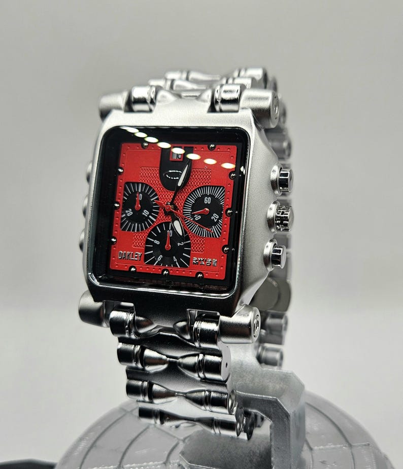 May include: A silver-toned Oakley watch with a square red face. The watch features multiple black dials and a silver metal band. The watch is displayed on a reflective surface.