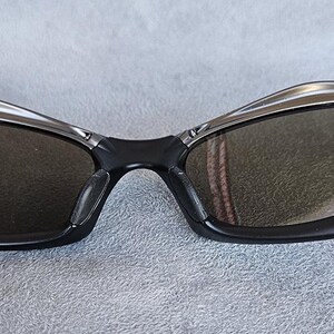 Puede incluir: Gafas de sol negras con un dise&ntilde;o elegante y moderno. Las monturas son negras con detalles plateados alrededor de las lentes. Las lentes son de color gris oscuro, proporcionando sombra y estilo. Un accesorio deportivo para actividades al aire libre.