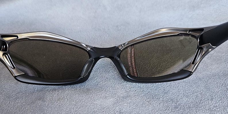 Puede incluir: Gafas de sol negras con un dise&ntilde;o elegante y moderno. Las monturas son negras con detalles plateados alrededor de las lentes. Las lentes son de color gris oscuro, proporcionando sombra y estilo. Un accesorio deportivo para actividades al aire libre.