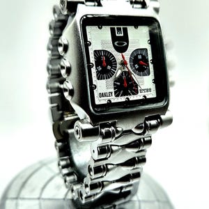 Pode incluir: Um rel&oacute;gio de pulso Oakley prateado com mostrador quadrado e pulseira de elos. O mostrador do rel&oacute;gio apresenta mostradores pretos e vermelhos e o logotipo Oakley. O rel&oacute;gio &eacute; exibido em um suporte esf&eacute;rico prateado.