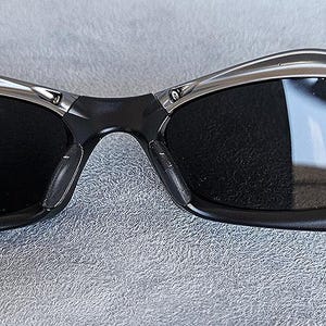 Peut inclure: Gros plan sur une paire de lunettes de soleil avec une monture noire et des verres fonc&eacute;s. Les lunettes de soleil ont un accent argent&eacute; sur le dessus et les c&ocirc;t&eacute;s de la monture. Les verres refl&egrave;tent l'environnement.