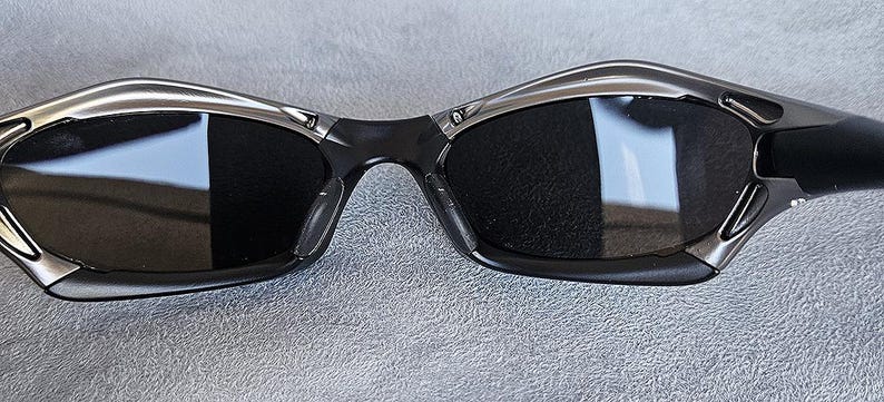 Peut inclure: Gros plan sur une paire de lunettes de soleil avec une monture noire et des verres fonc&eacute;s. Les lunettes de soleil ont un accent argent&eacute; sur le dessus et les c&ocirc;t&eacute;s de la monture. Les verres refl&egrave;tent l'environnement.