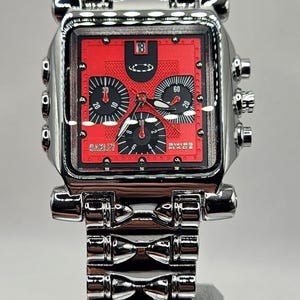 May include: A square-faced Oakley wristwatch with a red dial and silver-tone metal band. The watch face features black subdials and the number 18. The Oakley logo is visible. The watch band is made of multiple connected rectangular links.