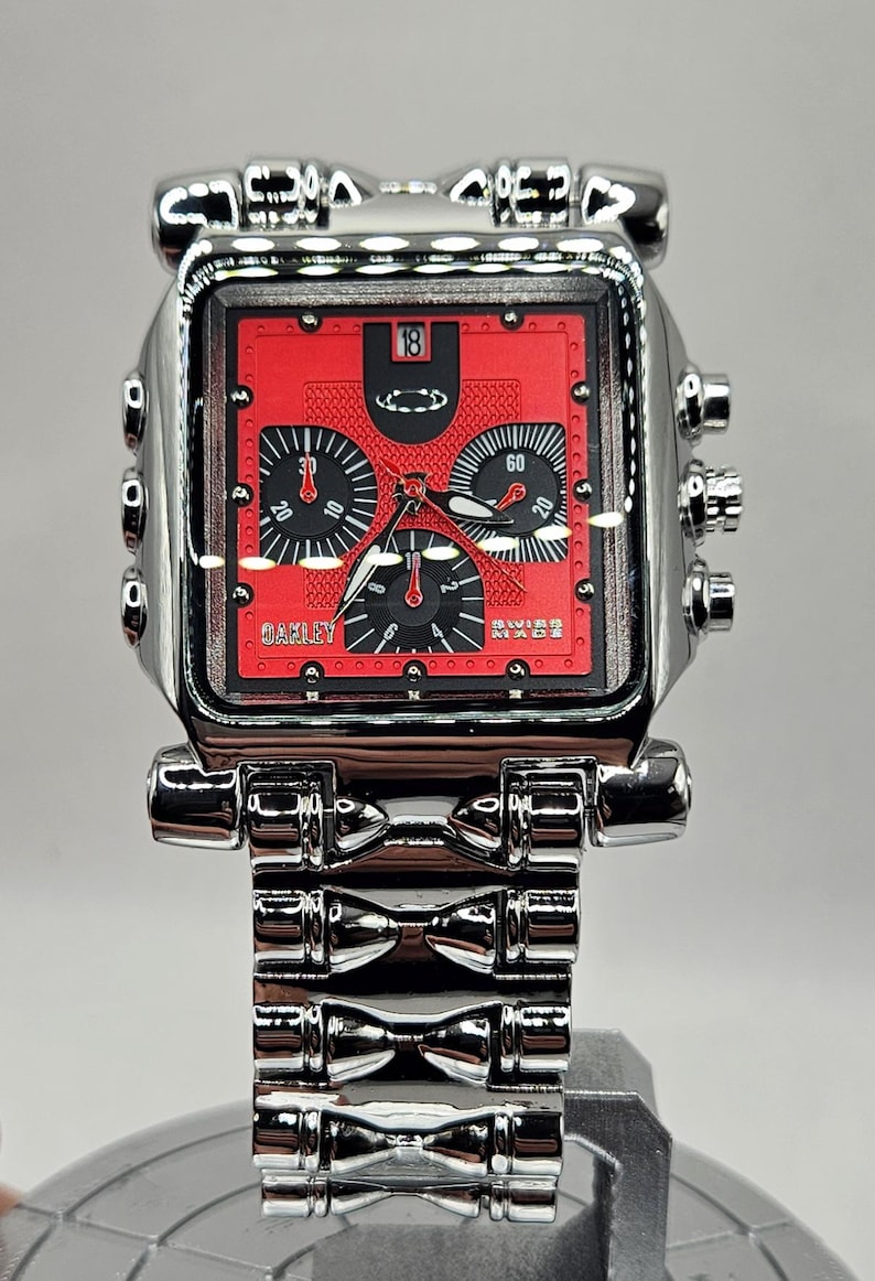 May include: A square-faced Oakley wristwatch with a red dial and silver-tone metal band. The watch face features black subdials and the number 18. The Oakley logo is visible. The watch band is made of multiple connected rectangular links.