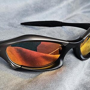 Puede incluir: Gafas de sol gris oscuro con lentes naranjas y amarillas. Las gafas tienen un dise&ntilde;o elegante y curvado con patillas negras y un peque&ntilde;o detalle de logotipo rojo. Las lentes reflejan el entorno.