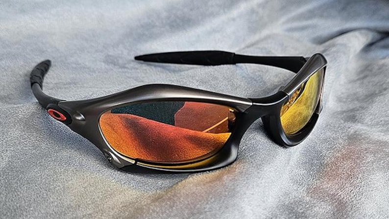 Puede incluir: Gafas de sol gris oscuro con lentes naranjas y amarillas. Las gafas tienen un dise&ntilde;o elegante y curvado con patillas negras y un peque&ntilde;o detalle de logotipo rojo. Las lentes reflejan el entorno.