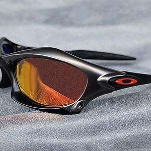 Puede incluir: Gafas de sol negras con lentes de color naranja. Las gafas tienen un dise&ntilde;o elegante y moderno con un acabado mate. El logotipo de Oakley es visible en rojo en las patillas. Se encuentran sobre una superficie gris.