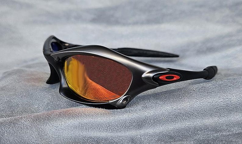 Puede incluir: Gafas de sol negras con lentes de color naranja. Las gafas tienen un dise&ntilde;o elegante y moderno con un acabado mate. El logotipo de Oakley es visible en rojo en las patillas. Se encuentran sobre una superficie gris.