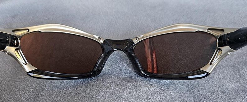 Może przedstawiać: Okulary przeciwsłoneczne z srebrną i czarną oprawką oraz brązowymi soczewkami. Okulary przeciwsłoneczne mają elegancki, nowoczesny design z zakrzywioną g&oacute;rą i czarnym mostkiem. Soczewki są w ciemnobrązowym kolorze.
