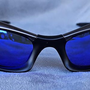 Puede incluir: Gafas de sol negras con lentes azul brillante. Las monturas tienen un dise&ntilde;o elegante y curvado, y las lentes reflejan el entorno. Las gafas de sol est&aacute;n sobre un fondo azul suave.