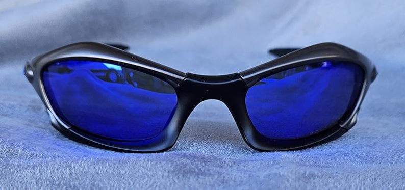 Puede incluir: Gafas de sol negras con lentes azul brillante. Las monturas tienen un dise&ntilde;o elegante y curvado, y las lentes reflejan el entorno. Las gafas de sol est&aacute;n sobre un fondo azul suave.