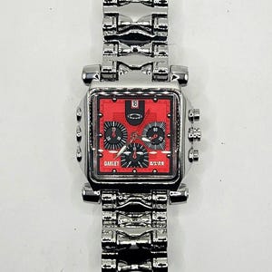 May include: A silver-toned Oakley wristwatch with a square red face. The watch features black subdials, a date display, and the Oakley logo. The watch band is made of linked silver metal.