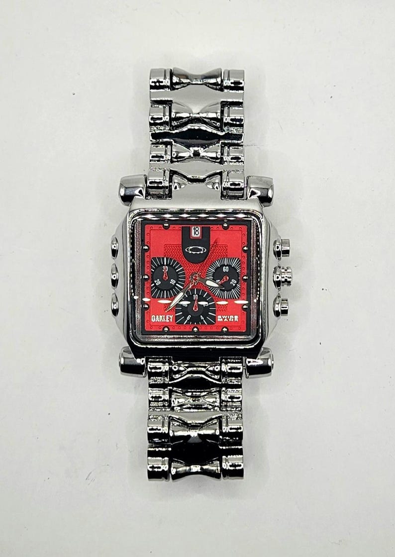 May include: A silver-toned Oakley wristwatch with a square red face. The watch features black subdials, a date display, and the Oakley logo. The watch band is made of linked silver metal.