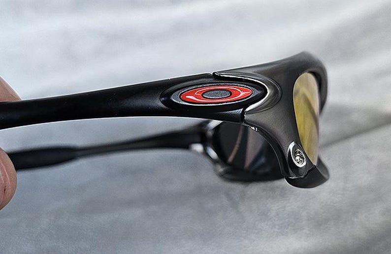 Puede incluir: Primer plano de gafas de sol negras con un logotipo rojo y blanco en la patilla. Las lentes son amarillas y la montura es negra mate. Las gafas de sol est&aacute;n en &aacute;ngulo, mostrando el lateral y la parte delantera.