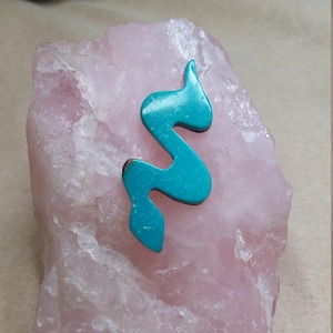 Könnte beinhalten: Ein türkisfarbener, abstrakter, schlangenförmiger Anhänger, der auf einem großen, rosafarbenen Rosenquarzkristall liegt.