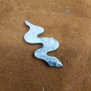 May include: A serpentine-shaped stone carving in shades of gray and white, with darker gray accents. The carving is set against a brown, textured background, showcasing the natural stone's unique patterns and form.