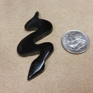 Może przedstawiać: Czarny, abstrakcyjny, falisty wisiorek z żywicy. Wisiorek jest mniej więcej wielkości polskiej dziesięciogroszówki.