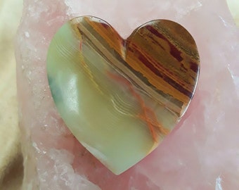 Puede incluir: Una piedra de ágata en forma de corazón de color verde, marrón y naranja sobre un fondo de cuarzo rosa.