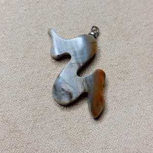 May include: A pendant made of a light brown, grey, and white stone with a silver bail. The stone is carved into a unique, abstract shape.