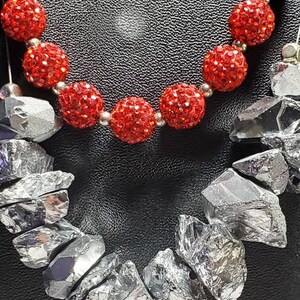 Puede incluir: Un collar con dos hileras de cuentas. La hilera superior está hecha de cuentas rojas brillantes. La hilera inferior está hecha de cuentas plateadas de forma irregular.