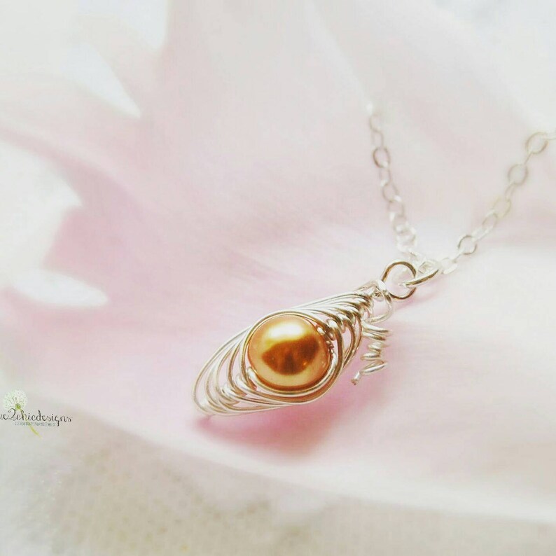 Pode incluir: Um colar de corrente de prata com um pingente que apresenta uma &uacute;nica p&eacute;rola dourada envolta em uma gaiola de arame. O pingente tem a forma de uma l&aacute;grima.