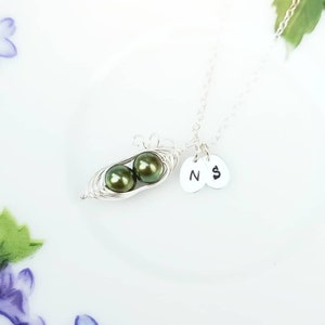 Pode incluir: Um colar de prata com um pingente de vagem de ervilha com duas p&eacute;rolas verdes. O colar tamb&eacute;m tem dois pingentes de prata com as letras 'N' e 'S' neles.
