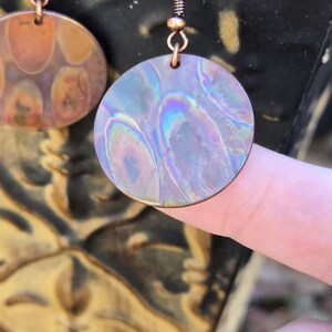 Pode incluir: Um par de brincos de cobre com um design redondo e iridescente. Os brincos t&ecirc;m uma textura martelada e um gancho de arame de cobre.