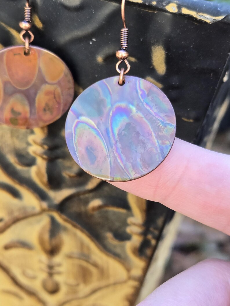 Pode incluir: Um par de brincos de cobre com um design redondo e iridescente. Os brincos t&ecirc;m uma textura martelada e um gancho de arame de cobre.