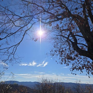 Pode incluir: Vista de uma cordilheira com um sol brilhante brilhando atrav&eacute;s dos galhos de uma &aacute;rvore nua em primeiro plano. O c&eacute;u &eacute; azul com algumas nuvens brancas.