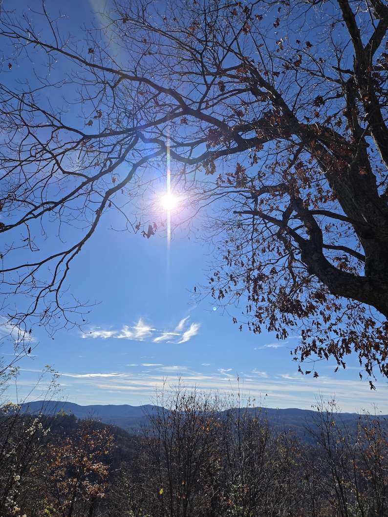 Pode incluir: Vista de uma cordilheira com um sol brilhante brilhando atrav&eacute;s dos galhos de uma &aacute;rvore nua em primeiro plano. O c&eacute;u &eacute; azul com algumas nuvens brancas.
