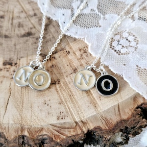 May include: Two silver necklaces with circular pendants spelling "NO". One pendant has a cream background with a silver "N" and "O". The other pendant has a black background with a silver "O". The necklaces are displayed on a wooden surface.