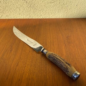 Op de afbeelding: Een vintage steakmes met een gebogen, getextureerd lemmet en een handvat van natuurlijk gekleurd geweihout. De handgreep is afgewerkt met zilverkleurig metaal. Het mes ligt op een houten oppervlak.