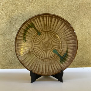 May include: A round, ceramic plate with a brown and tan radial design and green accents. The plate is displayed on a black stand against a textured, light-yellow background. The plate's design features a series of radiating lines from the center.