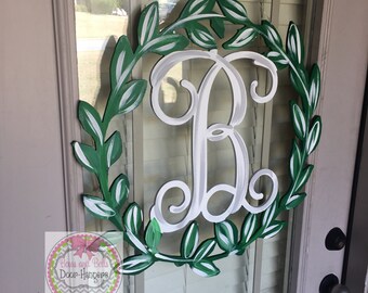 May include: A white wooden monogram letter "B" surrounded by a green wreath with white leaves. The wreath is painted on a wooden door hanger. The text "Bows and Bells Door Hangers" is visible in the lower left corner of the image.