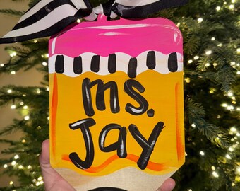 May include: A yellow and pink pencil-shaped wooden sign with black stripes and a black bow. The sign says "Ms. Jay" in black lettering.