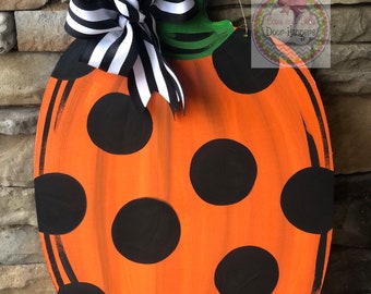 May include: Orange pumpkin door hanger with black polka dots and a black and white striped bow. The hanger has a green stem and is decorated with the text "Boos and Bells Door Hangers".