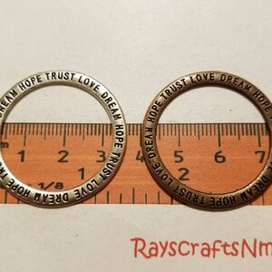 Puede incluir: Dos anillos metálicos con las palabras "Dream, Hope, Trust, Love" grabadas. Uno es plateado, el otro bronce. Una regla con medidas en pulgadas está en el fondo. Los anillos miden aproximadamente 2,5 cm de diámetro.