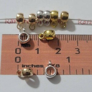 May include: A close-up of eight silver, gold, and mixed metal jump rings with a loop on the top. The jump rings are approximately 1 inch in diameter and are measured against a ruler.