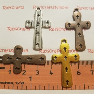 Puede incluir: Tres dijes de cruz de metal en diferentes acabados: plata, bronce y oro. Cada cruz tiene un diseño detallado con pequeñas cruces recortadas alrededor de los bordes. Los dijes se muestran en una regla con medidas en pulgadas.