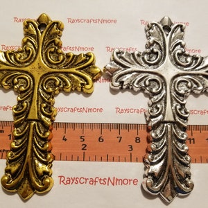 Puede incluir: Dos cruces de metal ornamentadas, una dorada y otra plateada, con intrincados adornos y un lazo en la parte superior. Las cruces miden aproximadamente 13 cm de largo. El texto "RaysCraftsNmore" es visible.