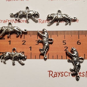 Puede incluir: Encantos de tigre de metal plateado con detalles de rayas y un lazo para sujetar. Una regla con medidas en centímetros es visible en el fondo. Ideal para hacer joyas o manualidades.