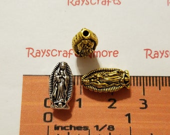 Puede incluir: Tres cuentas de metal plateadas y doradas con una imagen religiosa de la Virgen María. Las cuentas miden aproximadamente 2,5 cm de largo y se muestran junto a una regla.