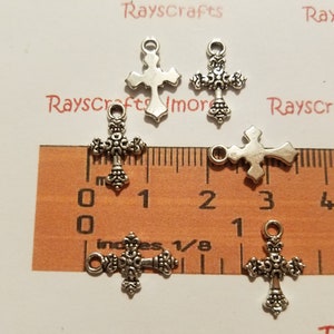 Puede incluir: Cinco dijes de cruz plateados con detalles ornamentados. Los dijes miden aproximadamente 2,5 cm de largo y se muestran en una regla.