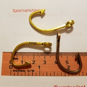 May include: Three metal fish hook charms in gold and copper tones. One gold hook has the word "HOPE" engraved on it. The hooks are approximately 1 inch long. A ruler is in the foreground.