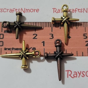 Puede incluir: Tres dijes de metal en forma de cruz con un diseño de clavo. Las cruces son en acabados dorado, bronce y cobre. Las cruces miden aproximadamente 2,5 cm de largo.